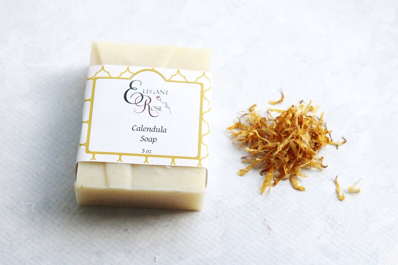 May include: A white bar of soap with a label that reads "Elegant Rose Calendula Soap 5 oz". Dried calendula petals are on a white surface next to the soap.