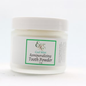 Op de afbeelding: Een witte pot Cool Mint Remineralizing Tooth Powder. De pot heeft een wit deksel en een etiket met groene randen en tekst. Het product is ontworpen voor mondhygiëne. Bevat 56,7 gram.