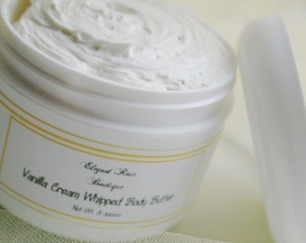 May include: A white jar of Vanilla Cream Whipped Body Butter from Elegant Rose Boutique. The jar is open, revealing a thick, creamy, white body butter. The label includes the product name and the net weight of 8 ounces.