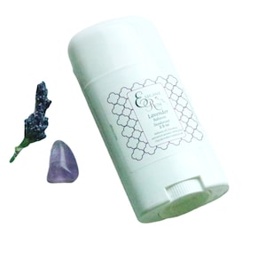 May include: White deodorant stick with a label that reads "Elegant & Rare" and "Lavender Balsamic Deodorant 2.5 oz". The label has a decorative pattern. There is a small purple flower and a purple crystal next to the deodorant stick.
