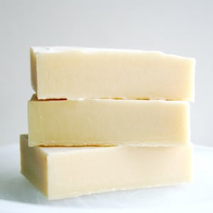 May include: Three rectangular bars of handmade soap stacked. The soap is a pale yellow colour and has a slightly rough texture. The bars are on a white plate. Each bar is approximately 10 cm long.