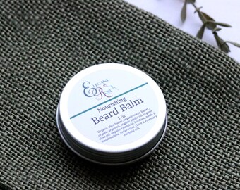 May include: A small silver tin with a white label that reads "Elegant Rose Nourishing Beard Balm 1 oz. Organic shea butter, organic cocoa butter, organic argan oil, organic jojoba oil, organic calendula, burdock, nettle & marshmallow, cedarwood, clove & rosemary essential oils"