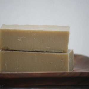 May include: Two rectangular bars of light beige soap are stacked on top of each other, resting on a wooden tray. The soap has a slightly rough texture and a natural appearance. The background is a soft, neutral color.
