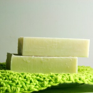 May include: Two bars of handmade soap, one on top of the other, are sitting on a green crocheted washcloth. The soap is a light green color and has a smooth, slightly textured surface.
