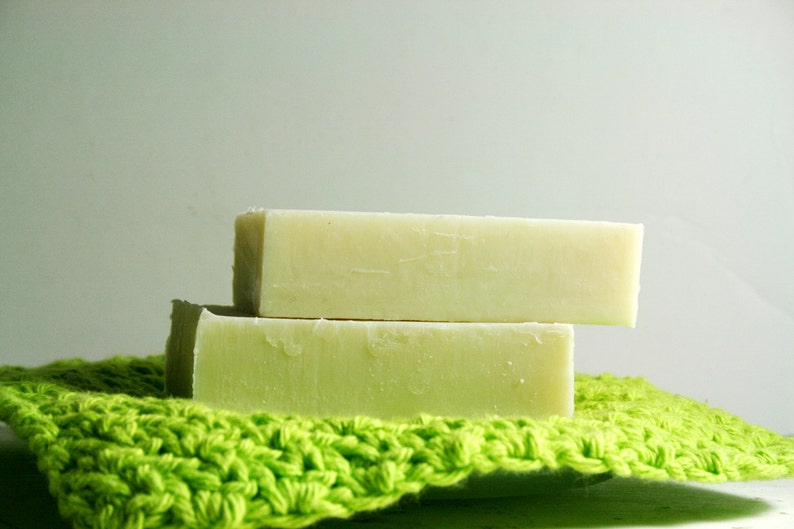 May include: Two bars of handmade soap, one on top of the other, are sitting on a green crocheted washcloth. The soap is a light green color and has a smooth, slightly textured surface.
