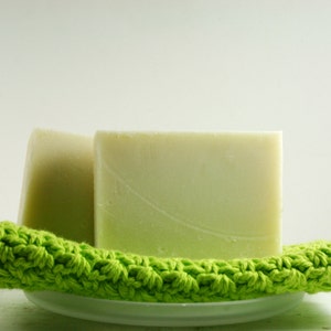May include: Two bars of handmade soap on a green crocheted dishcloth. The soap is a light green color and has a smooth, polished surface.