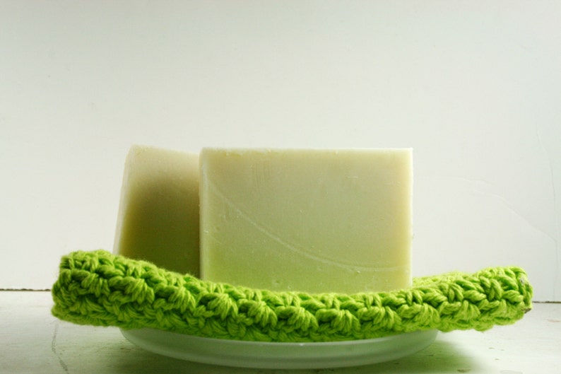 May include: Two bars of handmade soap on a green crocheted dishcloth. The soap is a light green color and has a smooth, polished surface.