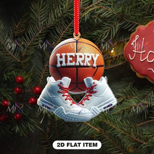 May include: A basketball-themed Christmas ornament featuring a basketball with the name "HERRY" and a pair of white sneakers with red laces. The ornament hangs from a red ribbon against a backdrop of pine branches.