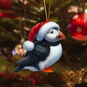 May include: A Christmas ornament featuring a puffin wearing a Santa hat. The bird has black and white feathers, an orange beak and feet, and a black scarf. The ornament hangs from a gold string against a blurred Christmas tree background.