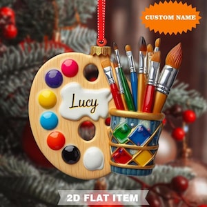 May include: A wooden artist's palette Christmas ornament with colorful paint splotches and a collection of paintbrushes. The name "Lucy" is written on the palette. The ornament has a red ribbon for hanging. Text on the ornament reads "CUSTOM NAME" and "2D FLAT ITEM."