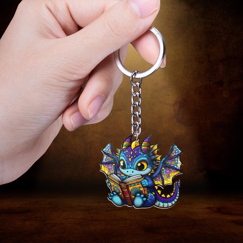 May include: A colorful dragon keychain with a silver chain and ring. The dragon is depicted reading a book, with blue, purple, yellow, and orange hues. The keychain is held by a hand against a brown background.