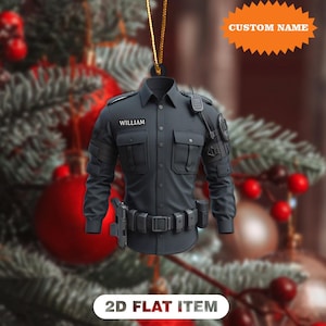 May include: A black police uniform ornament with the name "WILLIAM" on a name tag. The ornament is a 2D flat item and includes a gold string for hanging. The background is a blurred Christmas tree with red ornaments.