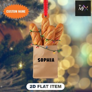 Può includere: Un ornamento natalizio con quattro baguette in un sacchetto di carta marrone, decorato con luci colorate. La borsa è personalizzata con il nome "SOPHIA". L'ornamento è appeso a un nastro rosso.