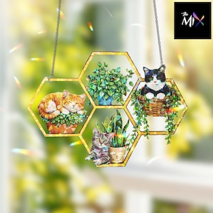 Puede incluir: Adorno decorativo colgante de gatos y plantas. El adorno presenta un diseño de panal con borde dorado, mostrando varios gatos y plantas en macetas. Los gatos están en diferentes poses y las plantas en macetas coloridas. El adorno cuelga de una cadena.