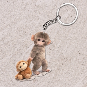Pode incluir: Um chaveiro com um design de macaco. O chaveiro mostra um macaco castanho claro segurando a mão de um macaco de pelúcia menor. Possui uma corrente e um anel prateados.