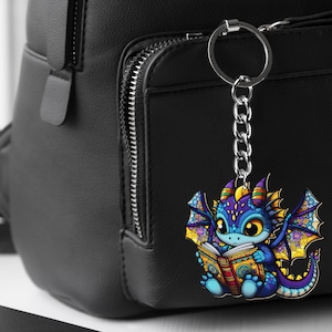 May include: A colorful dragon keychain with a silver chain and ring. The dragon is blue, purple, and yellow, reading a book with gold accents. The keychain is hanging on a black backpack.