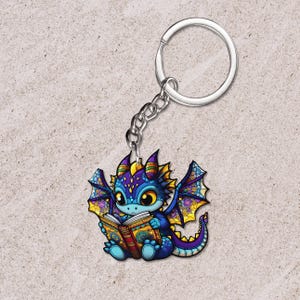 May include: A colorful dragon keychain with a silver chain and ring. The dragon is depicted reading a book, with blue, purple, yellow, and orange hues. The dragon has large eyes and detailed wings. The keychain is a fantasy-themed accessory.