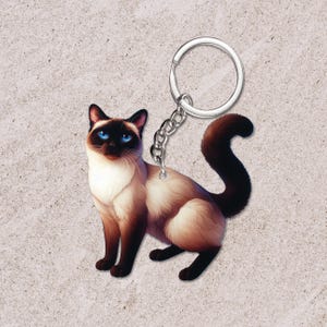 May include: A keychain featuring a Siamese cat with blue eyes. The cat is depicted in a sitting position with its tail curled around its body. The keychain is made of metal and has a silver finish.