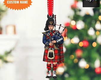 May include: A 2D flat ornament depicting a Scottish bagpiper in traditional dress. The ornament shows a bagpiper in a red and blue plaid kilt, a blue jacket, and a black hat. The name "DANIEL" is on the bag. It hangs from a red ribbon with white polka dots. The text "CUSTOM NAME" and "2D FLAT ITEM" are also visible.