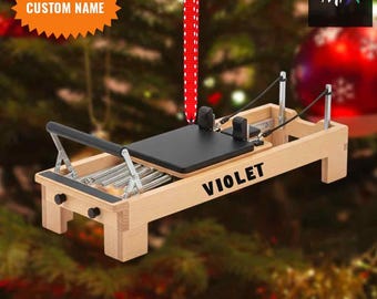 Könnte beinhalten: Holz-Pilates-Reformer-Ornament mit schwarzer Plattform und dem Namen "VIOLET". Es hängt an einem rot-weiß gestreiften Band. Die Texte "CUSTOM NAME" und "2D FLAT ITEM" sind ebenfalls sichtbar.