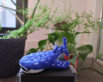 May include: A blue and white felt whale with white spots. The whale is lying on a black surface.