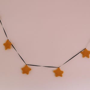 May include: A garland of five felt stars strung on black ribbon. The stars are a light yellow colour.