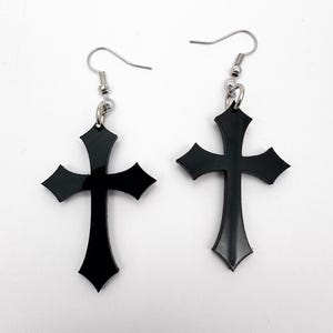May include: A pair of black cross earrings with silver-colored fish hook ear wires. The crosses have a gothic design with pointed edges. The earrings are displayed against a white background.