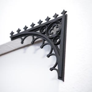 May include: A black, ornate metal bracket with a gothic design. The bracket is mounted on a white wall and features decorative cutouts and pointed finials. The bracket is a corner piece.