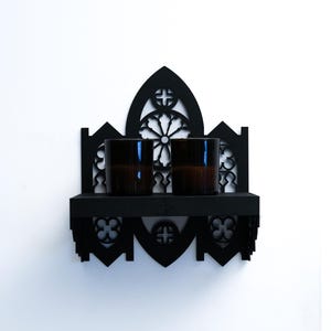 Peut inclure: Une &eacute;tag&egrave;re murale noire de style gothique, avec un sommet en arc et une petite &eacute;tag&egrave;re. Deux verres bruns sont pos&eacute;s sur l'&eacute;tag&egrave;re. Le design pr&eacute;sente des croix d&eacute;coup&eacute;es et des motifs floraux.