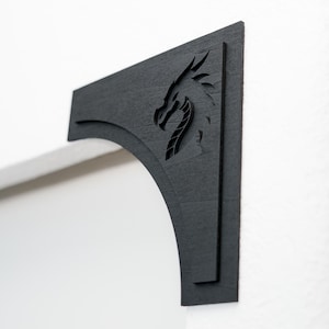 May include: A black wooden bracket with a dragon silhouette carved into the design. The bracket is mounted on a white wall, showcasing the dragon's head and neck. The bracket has an arched bottom and a right-angled top.