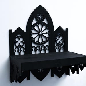 Peut inclure: &Eacute;tag&egrave;re murale noire de style gothique avec des motifs d&eacute;coup&eacute;s complexes. L'&eacute;tag&egrave;re pr&eacute;sente un panneau central en forme d'arche avec un motif circulaire et une croix, flanqu&eacute; de panneaux plus petits. L'&eacute;tag&egrave;re est rectangulaire et fix&eacute;e &agrave; un mur blanc.