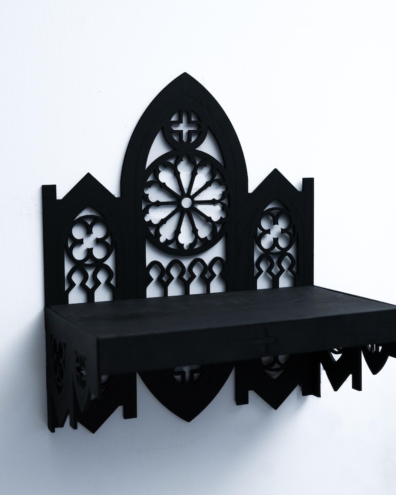 Peut inclure: &Eacute;tag&egrave;re murale noire de style gothique avec des motifs d&eacute;coup&eacute;s complexes. L'&eacute;tag&egrave;re pr&eacute;sente un panneau central en forme d'arche avec un motif circulaire et une croix, flanqu&eacute; de panneaux plus petits. L'&eacute;tag&egrave;re est rectangulaire et fix&eacute;e &agrave; un mur blanc.
