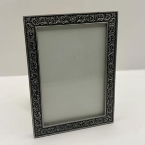 May include: A rectangular picture frame with a dark gray metallic finish. The frame features an intricate, raised relief pattern of scrolling vines, leaves, and small flower motifs along the entire border, surrounding a plain, light gray center.