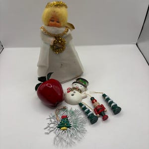 May include: A collection of holiday ornaments including a doll with yellow hair and a white gown, a red apple, a snowman, a nutcracker, two green and white striped cone-shaped ornaments, and a silver-coloured snowflake with a small tree centre.