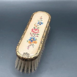 May include: An antique vanity brush with a cream-coloured top featuring a floral design in pink, blue, and yellow. The brush has a gold-coloured metal frame with ornate detailing and white bristles. A vintage beauty accessory.