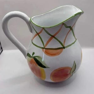 May include: A white ceramic pitcher featuring a hand-painted pattern of orange peaches with green leaves and a decorative green and orange lattice design around the upper rim. The pitcher has a curved handle and a wide, shaped pouring spout.