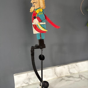 May include: A decorative nutcracker figure mounted on a black metal stand. The wooden nutcracker features a teal hat, a red and teal uniform with yellow accents, and a curved red lever. The figure is attached to a vertical black metal rod.