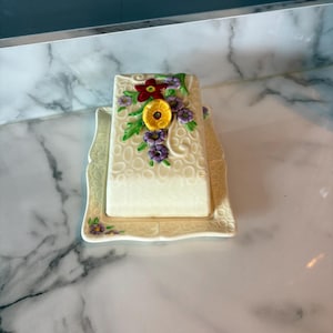 May include: A rectangular ceramic butter dish with a cream-colored base featuring a raised, embossed oval pattern. The lid is decorated with a cluster of hand-painted flowers in red, yellow, and purple, with green leaves and swirling vine accents.