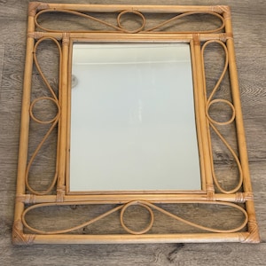 May include: A rectangular wall mirror featuring a light brown natural rattan frame. The frame is constructed with straight outer edges and decorative curved rattan loops that form symmetrical patterns along the top, bottom, and side borders.