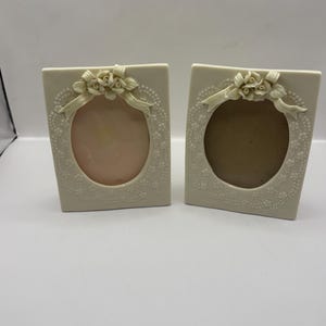 May include: Two off-white rectangular picture frames with oval openings. Each frame features a raised relief design of a floral bouquet with a ribbon at the top and a delicate dotted lace-like pattern surrounding the central oval display area.