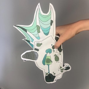 May include: A hand holding a cut-out illustration of a stylized creature. The creature is primarily white with green, blue, and black accents. The wings are a gradient of green and blue. The illustration has a whimsical, cartoonish style.