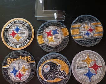 May include: Six round coasters with a Pittsburgh Steelers theme. Each coaster features a different design with rhinestones, including the team logo, a helmet, and the word "Steelers".