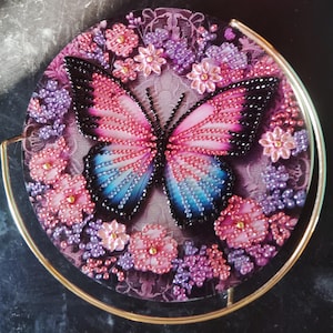 Op de afbeelding: Een rond decoratief stuk met een vlinderontwerp. De vlinder heeft roze, blauwe en zwarte kralen vleugels, omringd door roze en paarse kralen bloemen en bladeren. Een goudkleurig metalen frame omringt het ontwerp.