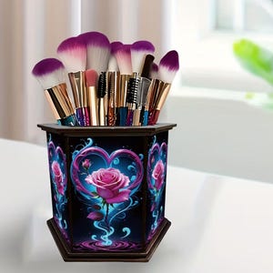 May include: A hexagonal makeup brush holder featuring a dark background with a vibrant pink rose centered inside a glowing blue heart graphic. The container is filled with various makeup brushes that have gold-toned ferrules and purple-tipped bristles.