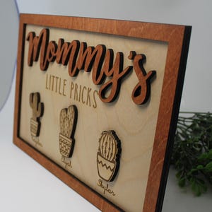 May include: A wooden sign with a brown frame and the text "Mommy's Little Pricks" in cursive. There are three cactus cutouts with the names "Cayden", "Elyjah", and "Skylar" underneath.