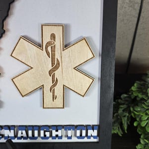 May include: A wooden framed sign with a black and white striped background. The sign features a wooden Star of Life with a snake and rod symbol. The name "MCNAUGHTON" is displayed in blue letters at the bottom of the sign.