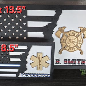 May include: Two wooden American flag shadow boxes with a firefighter and EMT emblem. The larger shadow box is 24 inches by 13.5 inches and the smaller shadow box is 15 inches by 8.5 inches. The firefighter emblem has a red fire helmet and the text "Fire Dept." The EMT emblem has a snake wrapped around a staff. Both shadow boxes have a black background and a white and red American flag design.