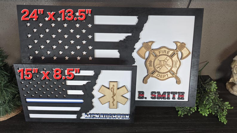 May include: Two wooden American flag shadow boxes with a firefighter and EMT emblem. The larger shadow box is 24 inches by 13.5 inches and the smaller shadow box is 15 inches by 8.5 inches. The firefighter emblem has a red fire helmet and the text "Fire Dept." The EMT emblem has a snake wrapped around a staff. Both shadow boxes have a black background and a white and red American flag design.