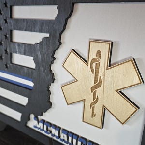 May include: A wooden framed shadow box with a black and white American flag, a blue police stripe, and a wooden Star of Life emblem. The text "MCMXCIX" is visible at the bottom of the frame.