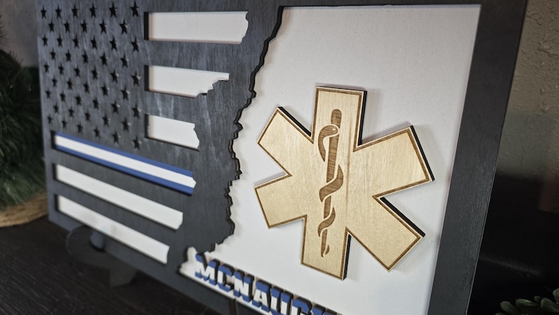 May include: A wooden framed shadow box with a black and white American flag, a blue police stripe, and a wooden Star of Life emblem. The text "MCMXCIX" is visible at the bottom of the frame.